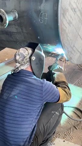 A person wearing a camouflage bandana, a dark blue striped shirt, black pants, and welding gear is kneeling and welding a large metal tank. Sparks are flying from the welding torch as the person works on the tank's exterior. The tank is marked with the number 67. The environment appears to be a workshop or industrial setting.