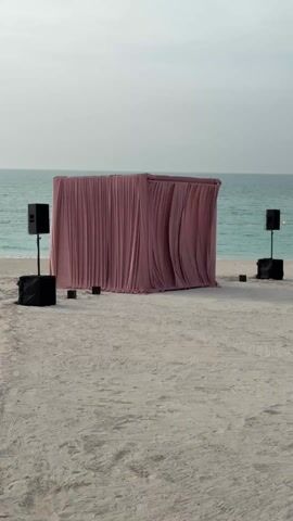 The scene is set on a sandy beach with a calm, light blue ocean in the background and an overcast sky. The central focus is a rectangular structure covered entirely in a dusty rose colored fabric, creating a closed-off space with draped curtains. Two black speakers on stands flank either side of the structure, and smaller black boxes are placed on the sand in front of them. There are no people visible in the frame.