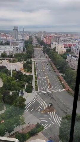 The video shows an aerial view of a wide, multi-lane road with traffic in a city. The road is lined with trees and buildings on both sides. There are pedestrian crossings with clear markings. The sky is overcast.