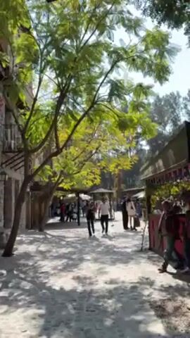 The video shows a sunny street scene with a cobblestone-paved path lined with buildings and trees. People are walking along the path in both directions. The buildings are mostly constructed of stone, and there are trees providing shade along the street. Some of the buildings appear to be shops or vendor stalls. There are various signs visible, including one that says "Stella". The lighting is bright, with shadows cast by the trees.