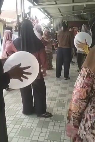 In the video, several individuals, predominantly women wearing headscarves, are gathered outdoors under a covered walkway. Many of them are holding white or polka-dotted balloons. One person in the center foreground is holding a large white balloon, and another person to their right is also holding a white balloon. To the left, a person in a black headscarf and maroon top is holding a white balloon. In the background, other individuals are visible, some of whom appear to be children. The environment seems to be a casual gathering or event. The floor is tiled, and the structure above suggests a roof or awning. The lighting indicates it is daytime. The overall mood appears festive or celebratory, given the presence of balloons and the gathering of people.