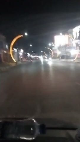 The video shows a nighttime view from a moving vehicle on a street. The street is lit by streetlights and decorative golden archway lights. There are other vehicles and motorcycles on the road. The interior of the vehicle is visible in the foreground, with a partially filled plastic water bottle on the dashboard. The scene appears to be in an urban or suburban area.