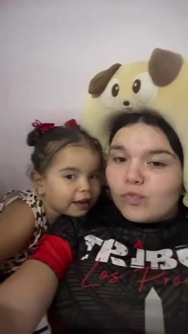 A selfie-style video shows a young girl and a young woman sitting closely together. The girl, with dark hair pulled up in a small bun adorned with a red bow, wears a sleeveless leopard print top. The woman, who appears to be slightly older, has long dark hair and is wearing a black t-shirt with red and white lettering and designs. Both individuals pucker their lips, creating a kissing face, and stick out their tongues for the camera. Behind them, a large, tan-colored stuffed animal in the shape of a dog with floppy brown ears is visible, leaning against what seems to be a plain white wall.