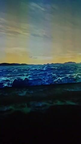 A dynamic projection displays a vast ocean under a twilight sky. The waves are a vibrant blue, with white crests that appear to be breaking. In the distance, silhouetted islands are visible against a warm, orange and yellow horizon. The sky transitions from a deep blue at the top to softer hues near the horizon, with wispy clouds. The foreground is a dark, indistinct area, suggesting a beach or foreground element. The perspective is low, as if the viewer is standing very close to the water's edge.