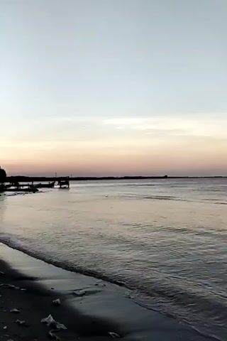 The video shows a serene beach scene at dusk. The camera pans across the water, revealing gentle waves lapping at the sandy shore. In the distance, a silhouetted pier extends into the water, with a few indistinct figures and structures visible on it. The sky is a soft gradient of pale blue and pastel pink, indicating the sun has recently set or is about to rise. The overall atmosphere is peaceful and tranquil, with the natural beauty of the coastline and the fading light dominating the frame.