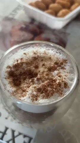 A close-up shot shows a glass of coffee topped with whipped cream and cinnamon. A spoon is used to stir the coffee, creating swirls in the cream. In the background, a container of pastries is visible. The scene is set on a table with a patterned tablecloth.