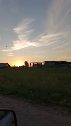 The video shows a sunset in a rural area. The sun is low on the horizon, casting a warm glow over the landscape. There are several houses and buildings scattered across the field, and a road in the foreground. The sky is filled with wispy clouds. A traffic sign for a pedestrian crossing is visible in the middle of the video. The video appears to have been filmed from a moving vehicle.