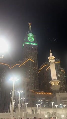 The video shows a nighttime view of the Makkah Royal Clock Tower, also known as Abraj Al-Bait, in Mecca, Saudi Arabia. The clock tower is illuminated with green lights, and an Arabic inscription is visible on its face. A minaret of a mosque is also visible to the right of the clock tower. The area below appears to be a plaza or courtyard, with many people gathered. Streetlights are also visible, casting a bright light on the scene. The sky is dark, indicating that it is nighttime. There are some faint lights in the sky above the clock tower, possibly birds or drones.