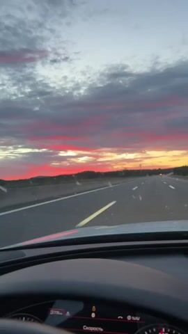 A first-person perspective video shows a sunset view from the driver's seat of a car on a highway. The sky is filled with dramatic clouds in shades of pink, red, orange, and gray. The highway is paved and has white lane markings. The car is in motion, indicated by the blur of the passing scenery and the dynamic perspective. The dashboard of the car is partially visible at the bottom of the frame, displaying some illuminated symbols. The overall scene captures the vibrant colors of a sunset experienced during a road trip.