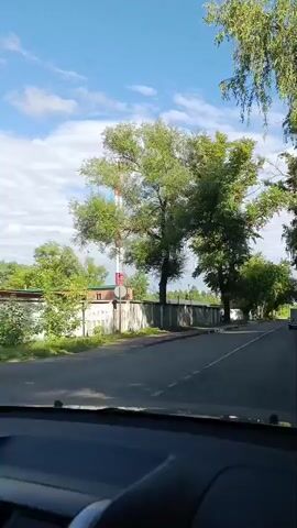 The video shows a drive-through scene on a sunny day in Orekhovo-Zuyevo, Russia. The camera perspective is from inside a vehicle, looking forward through the windshield. The road is paved and appears to be a two-lane street with a white line marking the center.

Along the right side of the road, there is a gray concrete fence, with trees and foliage visible behind it. On the left side of the road, a similar concrete fence runs alongside, with trees and some industrial structures, including a tall red and white tower, behind it. Further down, a large pile of cut logs is visible next to a building.

The sky is a vibrant blue with scattered white clouds, indicating good weather. Lush green trees line the sides of the road, with their branches forming a canopy overhead in some sections, creating a shaded, tunnel-like effect. The time of day appears to be daytime, likely late morning or early afternoon, given the bright sunlight.

A few vehicles are visible: a white van is ahead on the road, and another vehicle is partially visible further in the distance on the right. No people are clearly visible in the video. The overall mood is serene and typical of a drive through a semi-urban or industrial area with green spaces. No visible text is present in the video.