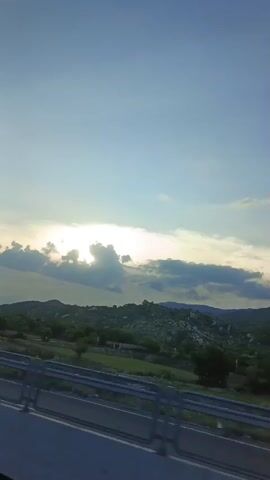 The video shows a landscape with rolling hills and a bright sky with clouds. The sun is visible behind the clouds, creating a light glow. In the foreground, there is a guardrail along what appears to be a road or highway. The vegetation on the hills is green and bushy. The overall impression is a scenic view from a moving vehicle.