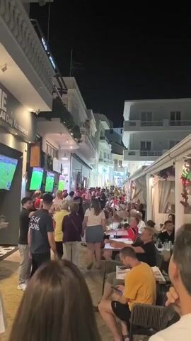 The video captures a lively outdoor scene at night on a narrow street lined with buildings and businesses. Numerous people are gathered, some standing and watching large screens mounted on the side of a building, while others are seated at tables outside of restaurants and bars. The atmosphere appears festive and energetic, with many individuals interacting and enjoying themselves. The screens show what seem to be sporting events. The street is illuminated by artificial lights from the businesses and overhead lighting.
