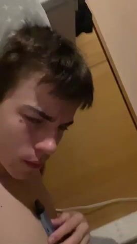A close-up video shows a young person lying down, with their head tilted. They are holding a blue vaping device near their mouth and appear to be inhaling or exhaling vapor. The person has dark, messy hair and their expression changes from serious to surprised. The background is blurry and appears to be a bed.