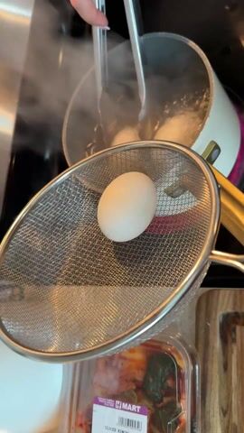 A person uses tongs to remove a white egg from a pot of boiling water on a stovetop. The egg is placed in a metal strainer held over a container of kimchi on a wooden cutting board. The tongs are then used to retrieve another egg from the pot. The stovetop is black with a red ring beneath the pot, which is white with a dark pink rim and gold-colored handles. Steam rises from the boiling water in the pot. Music plays in the background.