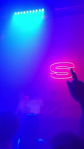 The video captures a dynamic scene inside a nightclub or music venue in Turin, Italy. Three young men, likely DJs or performers, are visible on a raised platform at the back of the room. One wears a white t-shirt with the text "SEXTAOY," while the others wear a white and a dark t-shirt respectively. Behind them, a prominent neon red "S" sign glows brightly on the wall. The lighting frequently shifts between intense red and a mix of purple and blue, creating an energetic ambiance. In front of the stage, a crowd of people, mostly seen in silhouette, are raising their hands, dancing, and cheering, indicating an engaging performance. The atmosphere is lively and exciting, typical of a night event.