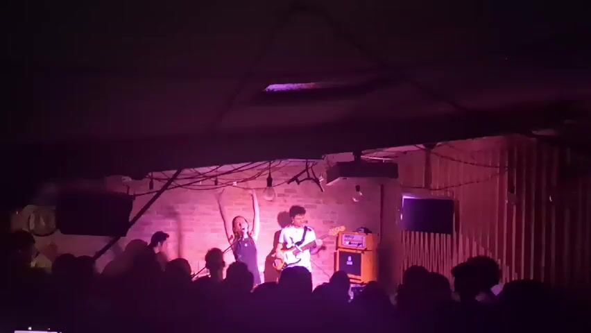 A band is performing on stage in a dimly lit venue. The band consists of at least four members. A female lead singer with long hair is standing center stage with her arms raised. To her right is a guitarist with curly hair, playing a white electric guitar. Behind them is a brick wall illuminated with pink light. An orange amplifier is beside the guitarist. Other band members are partially visible on the left side of the stage. A crowd of people is gathered in front of the stage, silhouetted against the light. There are wires and light fixtures hanging from the ceiling. There are also screens on either side of the stage.
