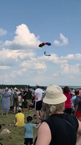 A skydiver descends under a colorful parachute, appearing to be performing a stunt with a long, dark fabric attached to their legs. Below, a large crowd of people are gathered on a grassy field, watching the skydiver. The crowd consists of individuals of various ages and genders, many of whom are casually dressed for an outdoor event. Some people are standing, while others are sitting or reclining on the grass. The environment is a bright, sunny day with a blue sky dotted with fluffy white clouds. The overall scene suggests an outdoor festival, airshow, or similar public gathering.