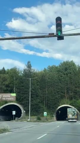 The video shows a view from a moving vehicle approaching and entering a tunnel. The scene is a highway surrounded by a dense forest of green trees. The sky is blue with scattered white clouds, suggesting it is daytime. There are two tunnel entrances visible, side-by-side. The left tunnel is dark and appears empty, while the right tunnel has a truck with a yellow cargo box just entering it. The truck's license plate is visible.

A green traffic sign with a white symbol of a tunnel entrance is visible on the right side of the road before the tunnel entrance. Another sign with text is partially visible on a billboard to the left of the road. The tunnel is arched with concrete supports and lined with lights. As the vehicle enters the tunnel, the interior is dark, illuminated by the tunnel lights and the truck's taillights.

The location is identified as Dolna Vasilitsa, Bulgaria. The overall mood is one of travel and transition, moving from an open, natural environment into an enclosed, man-made passage. The weather appears clear and pleasant. The activity is vehicular travel on a highway. There are no people clearly visible outside of the context of driving a vehicle. The prominent objects are the tunnels, the truck, the trees, and the road.