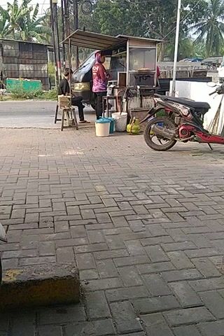 A red tent covers a food stall. A white truck is parked behind the stall. A red motorcycle is parked next to the stall. A person is sitting at the stall. The stall has a counter and shelves. There are pots and pans on the shelves. There are green gas tanks next to the stall. The stall is on a paved sidewalk. The sidewalk is made of gray bricks. There are trees in the background.