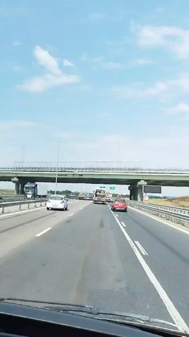 The video is taken from the perspective of someone inside a vehicle on a highway. The highway has multiple lanes and is covered by an overpass. There are other vehicles on the highway, including cars and trucks. The sky is blue with some clouds. The video appears to have been filmed during the day.