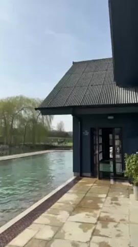 A building with a ridged roof and black-framed glass doors is next to a swimming pool. The building is dark gray, and the door is open. A stone patio with metal grating lies between the pool and the building. In the background are green trees and fields under a mostly sunny sky.