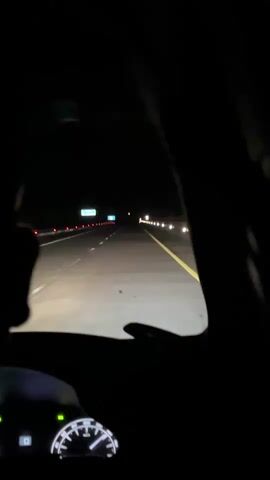 The video shows a point-of-view shot from inside a car driving on a highway at night. The headlights illuminate the road ahead, which has white lane markings and a yellow line on the right. Distant streetlights and signs are visible in the darkness. A dashboard with a speedometer is partially visible in the lower left corner. The scene is dark and the only light sources are the car's headlights and distant artificial lights.