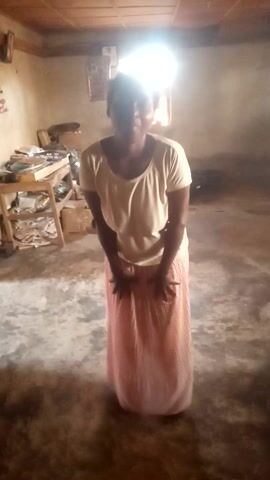 In a dimly lit room with bare walls and a concrete floor, a dark-skinned woman stands and poses for the camera. She is wearing a light-colored, possibly cream or beige, short-sleeved top and a long, pleated, reddish-brown or peach-colored skirt. A bright light source, likely a window, is visible on the left side of the frame, casting a glow and creating shadows.

In the background, a rustic wooden shelving unit or cart holds various items, though they are indistinct. The woman smiles brightly and playfully interacts with the camera, looking directly at it for most of the video. She moves her hands, at times holding or adjusting her skirt, and occasionally looks down or to the side before returning her gaze to the camera. Her demeanor suggests a happy and cheerful mood. The setting appears to be an indoor space in Gudige Sabo, Nigeria, with a humble and utilitarian aesthetic. There is no visible text.