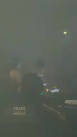 The video shows a DJ at a club or concert venue. The DJ is operating a Pioneer DJ setup, illuminated by flashing white lights. The environment is dark and smoky with a crowd of people visible in the background. There are signs reading "TRANS MUSICALES" at the bottom of the setup. The scene is characterized by pulsing light and the energy of a live music performance.