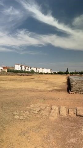 A vertical video shows an outdoor archaeological site under a bright sky with scattered clouds. The foreground features uneven dirt ground with some exposed stone blocks arranged in a line. To the right, a weathered, wooden, cube-shaped structure, possibly a protective cover or display, is situated on the dirt. In the middle ground, a low, rubble-like stone wall or foundation runs horizontally, with more exposed stone work beyond it. The background reveals a line of white buildings with red-tiled roofs, indicative of a town or city. Trees and vegetation are visible beyond the buildings and archaeological features. The overall impression is of an ancient ruin partially excavated and preserved within a modern landscape.