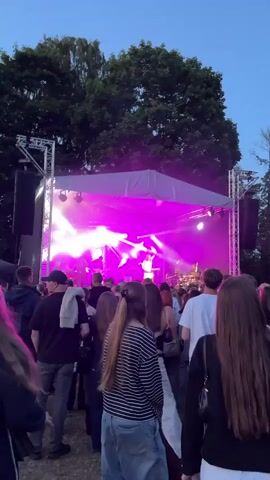 This is a vertical video of an outdoor concert at dusk in Panevėžys, Lithuania. A band is performing on a brightly lit stage with purple and white lights. A female singer in a light-colored dress is at the front of the stage, with other band members behind her. A crowd of people are gathered in front of the stage, watching the performance. The sky is a deep blue, indicating it's twilight. Large trees with dark green foliage surround the concert area. The overall atmosphere appears to be lively and engaging, with the audience focused on the performers. The video captures the energy of a live music event during the evening.