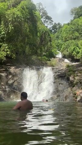A wide shot depicts a scene at a waterfall. A man with dark hair and shirtless, his back to the camera, is standing waist-deep in the water. A second person, with lighter hair, is visible further out, closer to the waterfall. Water cascades down the rocks in the center of the frame, forming a waterfall. Lush green vegetation surrounds the area, with trees and foliage covering the landscape. The sky is overcast, suggesting daylight conditions.