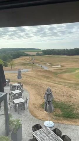 The video captures an outdoor scene with a golf course in the background and a patio with tables and chairs in the foreground. The golf course appears dry and parched, with rolling hills and a winding path. Two individuals are seen near a golf cart on the path. The sky is cloudy. The patio area is furnished with outdoor dining sets, some with umbrellas. The perspective suggests the video is taken from a higher vantage point, possibly from within a building overlooking the golf course.