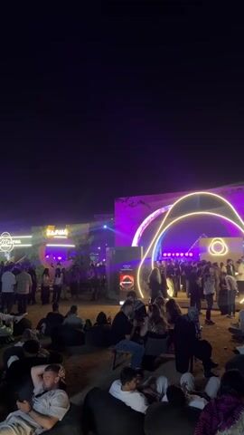 The video shows an outdoor nighttime scene at what appears to be a festival or event. There are many people milling around, some standing and talking, and others sitting in beanbag chairs in the foreground. In the background is a structure with the word “ZANA” displayed on it, illuminated in bright pink and purple lights. A yellow neon light feature in the shape of intersecting circles adds to the visual spectacle. A searchlight beams upwards into the dark night sky. The overall atmosphere suggests a lively, festive gathering.