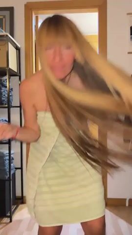 A person with long blonde hair, wearing a light green striped towel, is dancing energetically in a room. Their hair is being flung around dramatically as they move. The setting appears to be a home, possibly a bedroom or bathroom area, with a wooden door in the background. Shelving units filled with what look like folded towels or clothing are visible on the left side of the frame. A framed picture or artwork hangs on the wall behind the person. The lighting suggests it is daytime. The person's facial expression is joyful and animated, indicating a playful and lively mood.