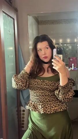 The video is a selfie video taken by a young woman in a bathroom. She is wearing a leopard print off-the-shoulder top with puffed sleeves and a green satin skirt. She has on a black choker necklace and a pink bracelet on her left wrist. The woman is holding her phone up to take the selfie and is touching her hair with her right hand. She is looking at her reflection in the mirror. In the background, there is a wooden door on the left, a laundry basket, a mirror with shelves above it that hold various toiletries and cosmetics, and a toilet with a trash can next to it. The lighting is bright, suggesting daylight.