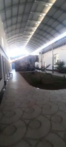 The video shows a long hallway with an arched, corrugated metal ceiling. On one side, there are multiple windows with black frames. On the other side, there is an open area with plants and a rock formation. The floor is covered in patterned tiles. The person filming is walking through the hallway.