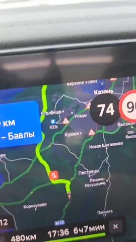 A first-person view from inside a car driving down a highway. The car's navigation system is visible, showing a map with the route highlighted and the car's speed at 75. The map indicates the destination as "Bavly." The dashboard is partially visible, and outside the car, the view shows a multi-lane highway with trees on either side. Several trucks are seen driving in the same direction as the car. The sky is mostly clear with some clouds.