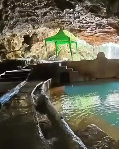 The video shows a natural pool of turquoise water inside a cave. The cave walls are rough and rocky, with some vegetation visible. In the center of the cave, there is a green tent set up next to a stone structure with steps leading down to the water. The water appears shallow and clear.