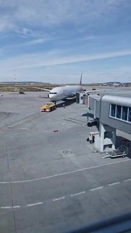 An aerial view captures an airport tarmac on a sunny day. A large passenger airplane, emblazoned with the "РОССИЯ" logo and a distinctive red and white pattern on its tail, is parked at a gate. A jet bridge extends from the terminal to the aircraft's door, and a yellow tug vehicle is positioned at the front landing gear, appearing to be connected for towing. A person in a high-visibility vest stands near the aircraft's nose. In the background, other airport structures, vehicles, and distant hills are visible under a blue sky with scattered clouds. The tarmac is marked with white lines and painted symbols, indicating the operational nature of the environment.