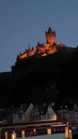 The video shows a castle illuminated by orange lights on a hill at dusk. Below the castle, there is a village with half-timbered houses. The sky is dark blue. In the foreground, there is a railing, suggesting the viewpoint is from a balcony or boat.