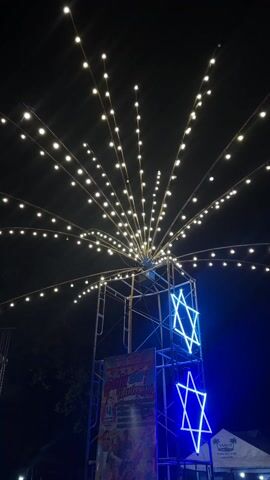 The video shows a nighttime scene featuring a tall metal structure adorned with illuminated Star of David neon signs. Above the structure, numerous strings of lights radiate outwards in a radial pattern. The structure is surrounded by a dark background, with the edge of a tent visible on the right side. A banner with text and images is attached to the structure. There is music playing in the background.