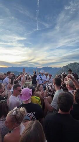 A crowd of people are gathered on what appears to be a mountaintop or elevated area overlooking a body of water and mountains in the distance. The sky is blue with dramatic clouds. Many people are raising their hands in the air, suggesting they are at a party or concert. In the center of the crowd, a person or people appear to be DJing or performing, with equipment visible. The overall atmosphere is one of celebration and excitement. The lighting suggests it is late afternoon or early evening.