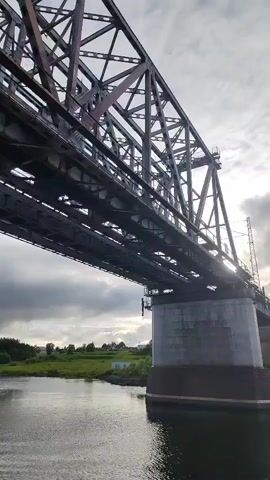 The camera is positioned below a large metal bridge, angled upwards. The bridge has a complex lattice structure and is supported by a concrete pillar in the water. The water appears calm and dark. On the shoreline opposite the pillar, there's a green grassy area, a few trees, and a small white building visible in the distance. The sky is overcast with gray clouds. The sunlight appears to be filtering through the clouds.