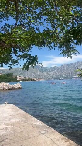 A video captures a scenic view of a bay with mountains in the background and a town nestled along the coastline. The water is a clear turquoise color, and the sun is shining brightly, creating a beautiful, serene atmosphere. Several small boats are visible on the water, and a large cruise ship is docked in the distance. The foreground shows a stone pathway, and lush green trees with leaves frame the top of the video. The overall impression is one of a peaceful and picturesque vacation destination.