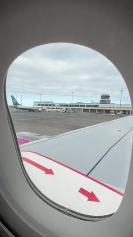 The video shows a view from an airplane window, looking out at the tarmac. Another Ryanair plane is parked at the terminal. The weather is overcast. Red arrows on the wing of the plane point towards the back of the plane.