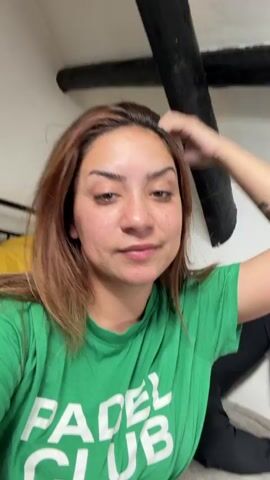 This is a short video showing a woman with brown hair and fair skin wearing a green t-shirt. She is looking at the camera and touching her hair. Her expression changes from a neutral look to a pout. In the background, there is a yellow object, a black punching bag, and part of a staircase with wooden beams.