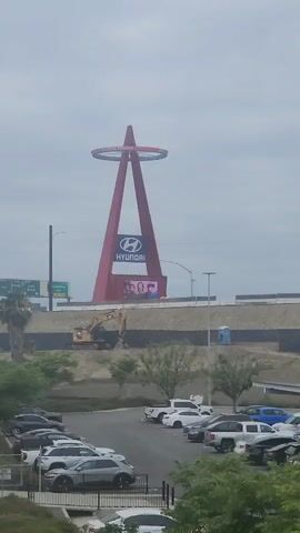 This media file captures a wide, outdoor scene with a prominent red A-frame structure topped with a halo-like ring, bearing the "ANGELS" logo. Below the ring, a large Hyundai logo is displayed, and a digital billboard shows an advertisement. The structure appears to be a landmark, possibly a stadium or monument.

In the foreground, a paved bike path runs alongside a dirt path. Several cyclists are visible on the bike path, moving from left to right in the frame. Further back, there's a parking lot filled with various vehicles, including sedans and SUVs.

Behind the bike path and parking lot, a construction zone is evident. An excavator is at work on a raised embankment, and a portable toilet is visible. There are trees and palm trees scattered throughout the scene, suggesting a warm climate. Highway signage indicating directions and exits is also present, including a sign for "STATE COLLEGE BLVD."

A "ROAD WORK AHEAD" sign is clearly visible on the right side of the frame, indicating ongoing construction or maintenance. The sky is overcast, suggesting a cloudy or hazy day. The overall atmosphere is utilitarian, with elements of urban infrastructure, recreational activity, and construction work. The visible text includes "ANGELS," "HYUNDAI," and directional highway signs. The time of day appears to be daytime, but the overcast conditions limit precise time estimation.