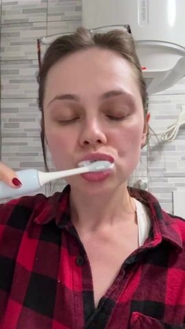 A woman is brushing her teeth with an electric toothbrush in a bathroom. She is wearing a red and black flannel shirt over a white tank top, and her hair is pulled back. She has on red nail polish and places glasses on her head in the later half of the video. She looks upwards as she brushes. The bathroom has light grey and white striped tiles and an Atlantic water heater on the wall.