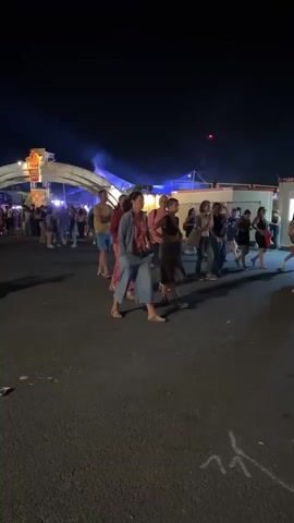 The video shows a nighttime outdoor event with a group of people walking in a line. There are colorful lights and structures in the background, suggesting a festival or fair. The people are dressed casually, and some are looking towards the camera. The ground is paved and appears to be a dark asphalt or concrete surface. There are some white markings on the ground, possibly for parking or directional purposes. The sky is dark, indicating it is nighttime. The overall atmosphere appears to be festive and lively.