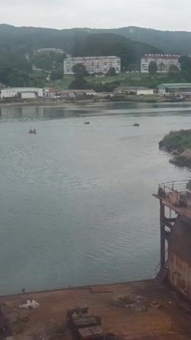 The video shows a wide shot of a body of water, possibly a river or bay, under an overcast sky. In the foreground, a portion of a rusty, dilapidated structure, likely a dock or a derelict ship, occupies the lower right corner and bottom of the frame. The water is a dull grey-green, with some ripples visible. Several small boats are scattered across the water. On the far bank, there is a town or settlement with buildings, including multi-story apartment complexes, nestled on a tree-covered hillside. The overall atmosphere is somewhat bleak and industrial.